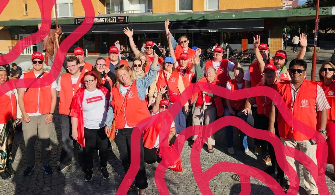 Noin 30 henkilöä katsoo kameraan ja vilkuttaa, osa siristää kirkkaan auringonvalon takia. Monet nauravat tai hymyilevät. Koko porukalla on päällä punaiset Ruotsin sosialidemokraattien liivit.